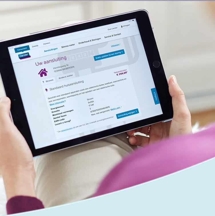 Een persoon bekijkt een tablet waarop een scherm te zien is met informatie over een elektriciteitsaansluiting op de website van Liander. Op het scherm staat "Uw aansluiting" en er worden details weergegeven, zoals het adres, de netwerkkosten, en kenmerken van de aansluiting, zoals energiebron, grootte, en maximale vermogen. De hand van de persoon is zichtbaar, en de achtergrond bestaat uit een lichte bank of stoel.