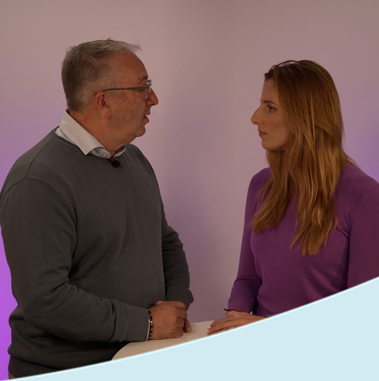 Twee personen zitten tegenover elkaar aan een ronde tafel en voeren een gesprek in een studio met paarse achtergrondverlichting