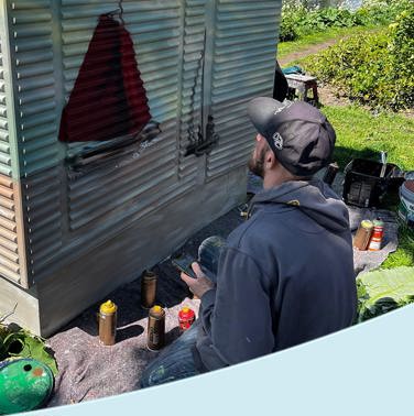 Een kunstenaar zit op zijn knieën in het gras en werkt aan een muurschildering op een metalen elektriciteitskastje aan het water. Op het kastje is een rode zeilboot in opbouw te zien. Rondom liggen verfspullen, spuitbussen en verfemmers. Op de achtergrond staan bomen en een aantal gebouwen langs de oever onder een strakblauwe lucht.
