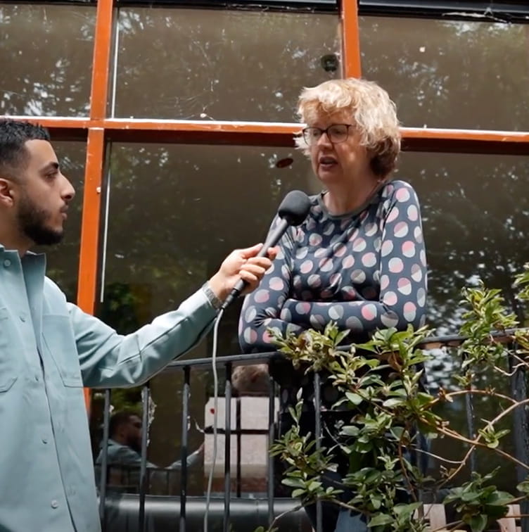 Man interviewt vrouw bij haar huis terwijl ze op haar balkon staat. Liander werkt in haar buurt aan het energienet. 