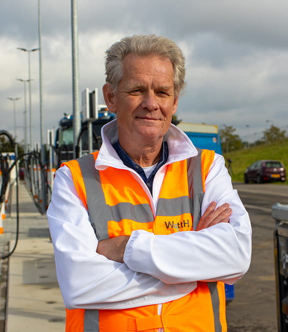 Gerlach van betuwewind staat met zijn armen over elkaar op het laadplein Betuwewind. Hij kijkt glimlachend de camera in, en draagt een wit hemd met daar overheen een oranje veiligheidshesje