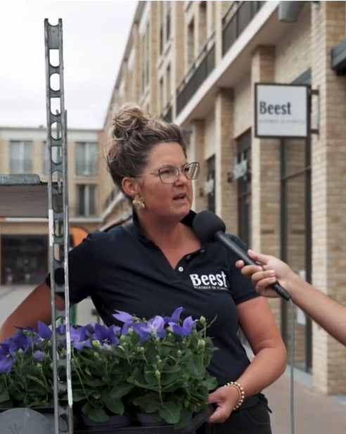 Een persoon staat in een woonstraat met een plantenbak in de hand terwijl er een microfoon wordt vastgehouden. Op de achtergrond zijn moderne woningen te zien.