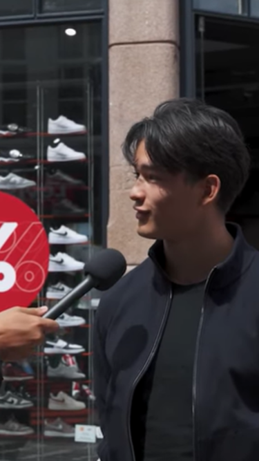 Twee mensen staan in een winkelstraat bij een etalage met schoenen, terwijl een microfoon wordt vastgehouden tijdens een straatinterview.