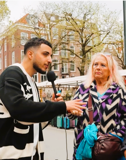 Interviewer in gesprek met een persoon op een marktplein, met kraampjes en gebouwen op de achtergrond.