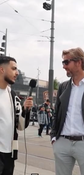Straatinterview op een druk plein waar een interviewer in gesprek is met een man in pak en zonnebril, omringd door voorbijgangers in een stedelijke omgeving.