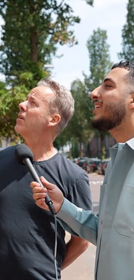 Interviewsituatie buiten waarbij iemand een microfoon vasthoudt en een ander persoon te woord staat, met bomen en een straat op de achtergrond.