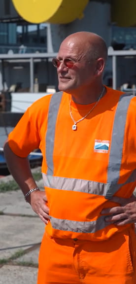 Persoon in oranje veiligheidskleding met reflecterende strepen op een industriële locatie.