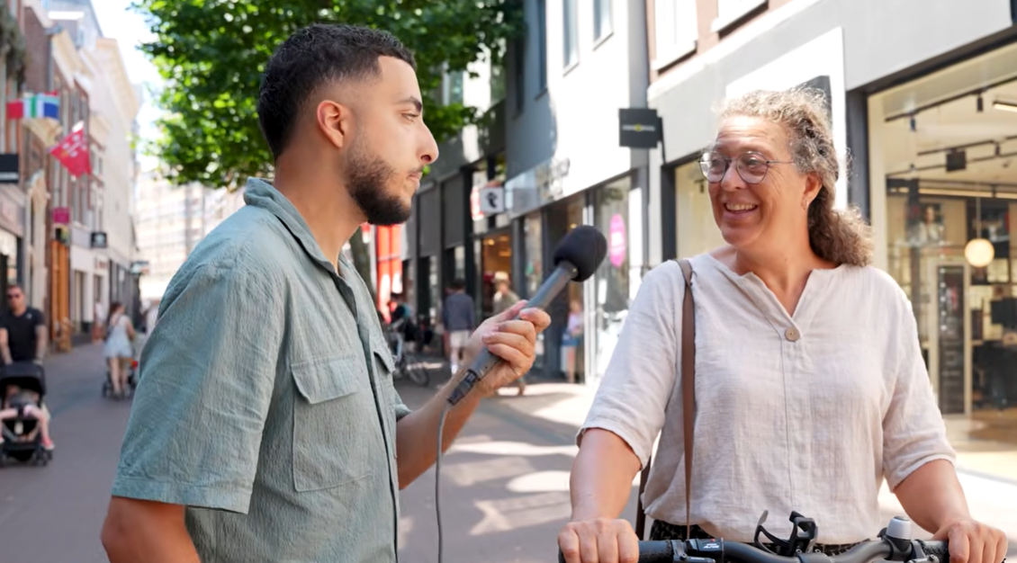 In een winkelstraat wordt een gesprek gevoerd terwijl iemand met een fiets stilstaat. De setting laat een alledaagse situatie in de stad zien, passend bij het thema slim omgaan met energie.