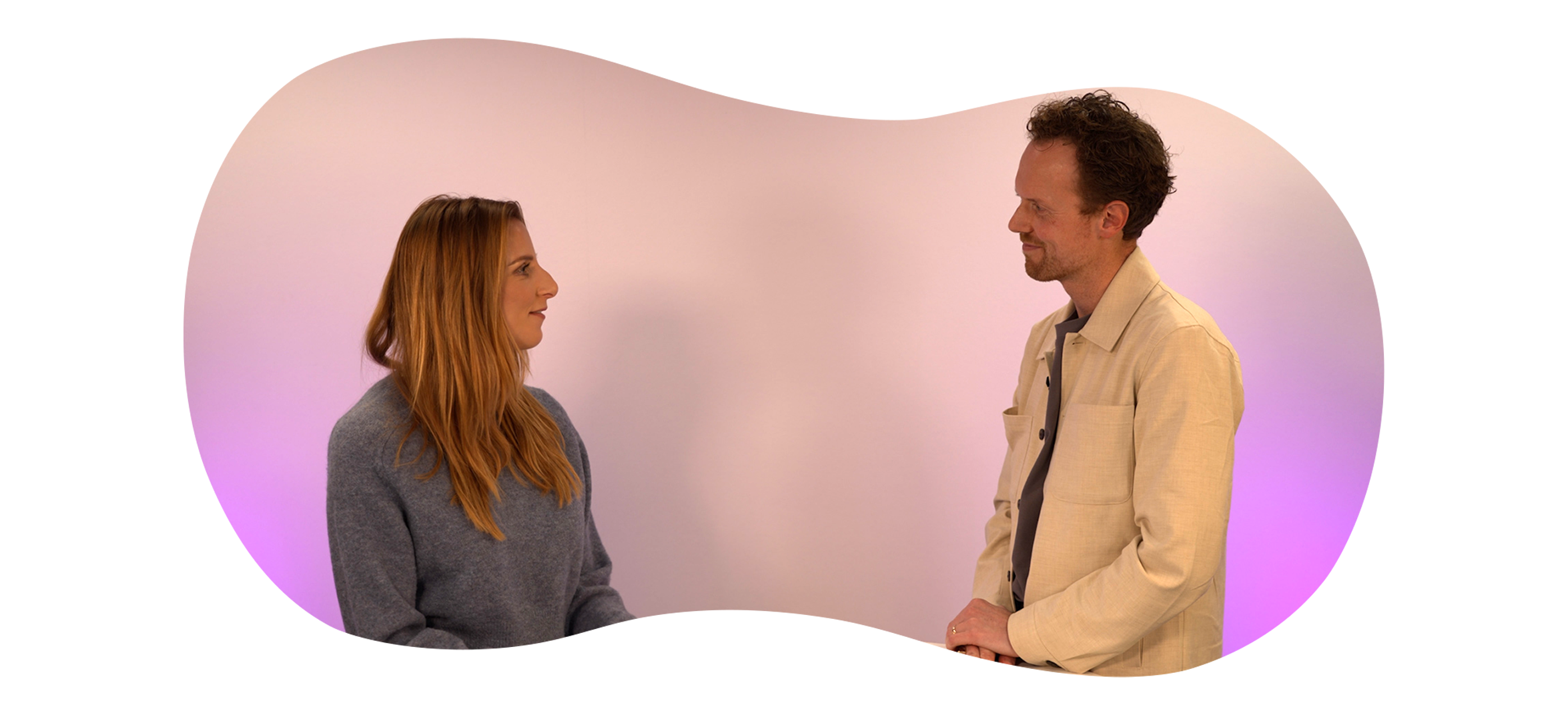 Twee personen staan tegenover elkaar en zijn in gesprek in een studio met een lichte achtergrond en paarse verlichting.