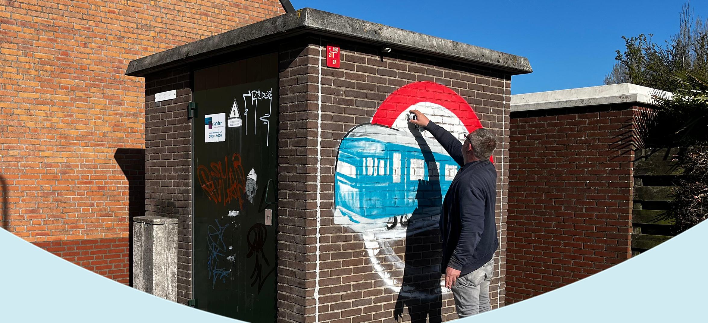 Een man schildert een muurschildering op een elektriciteitshuisje in een woonwijk. Op de muur is een afbeelding in aanbouw te zien van een blauwe trein in een rode cirkel. Naast hem liggen verfspullen en een blauwe deken op de stoep. Het is een zonnige dag met een strakblauwe lucht.