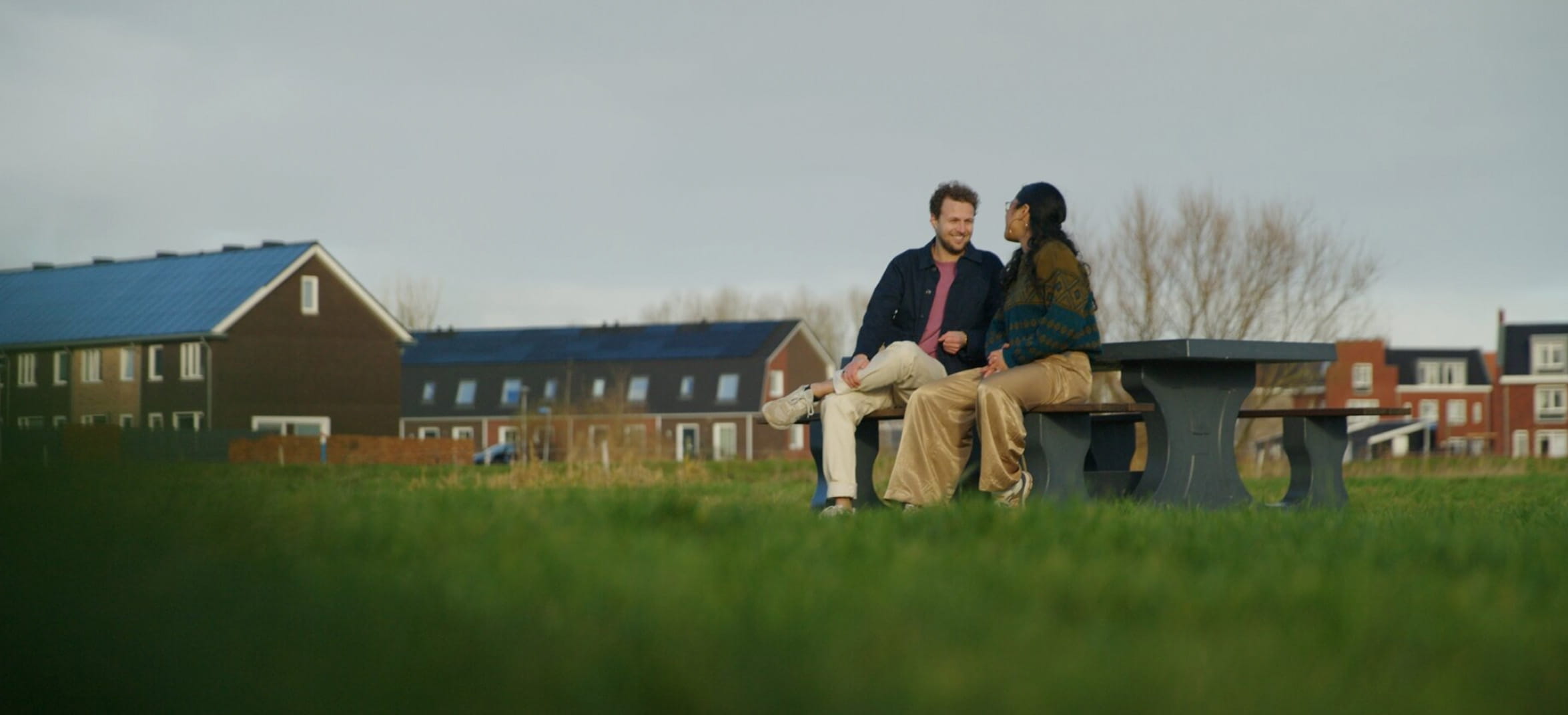 Een man en een vrouw zitten op een bankje in een groen landschap met achter hen huizen. Ze praten.