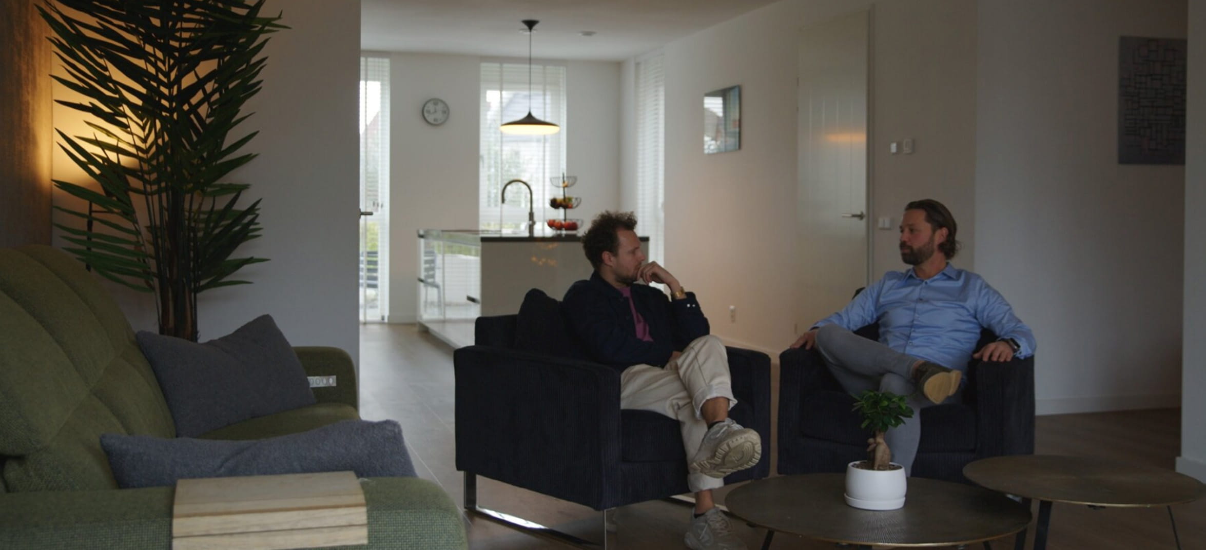 Twee mannen zitten naast elkaar op stoelen in een woonkamer en praten met elkaar.