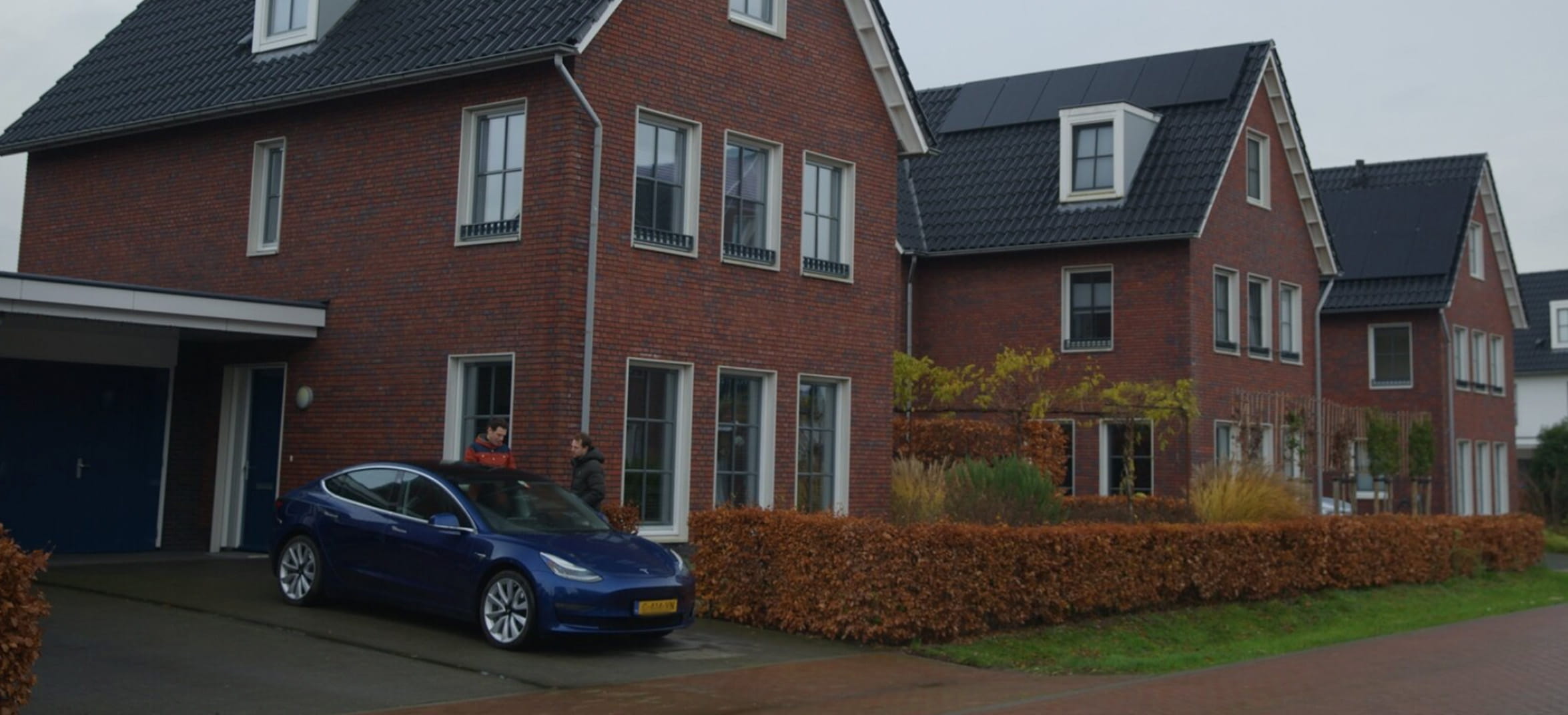 Twee mannen staan achter een tesla-auto bij een vrijstaand huis. Ze praten met elkaar over de thuislaadstekker.