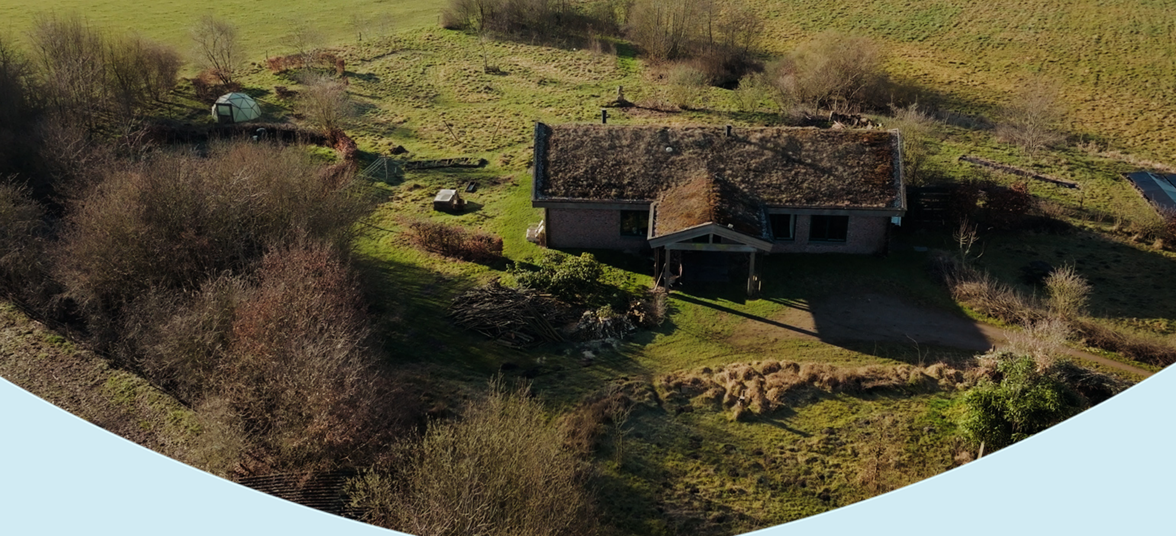 Landelijk huis met groen zedem dak omringd door natuur, struiken en een geodetische koepel in een open landschap.