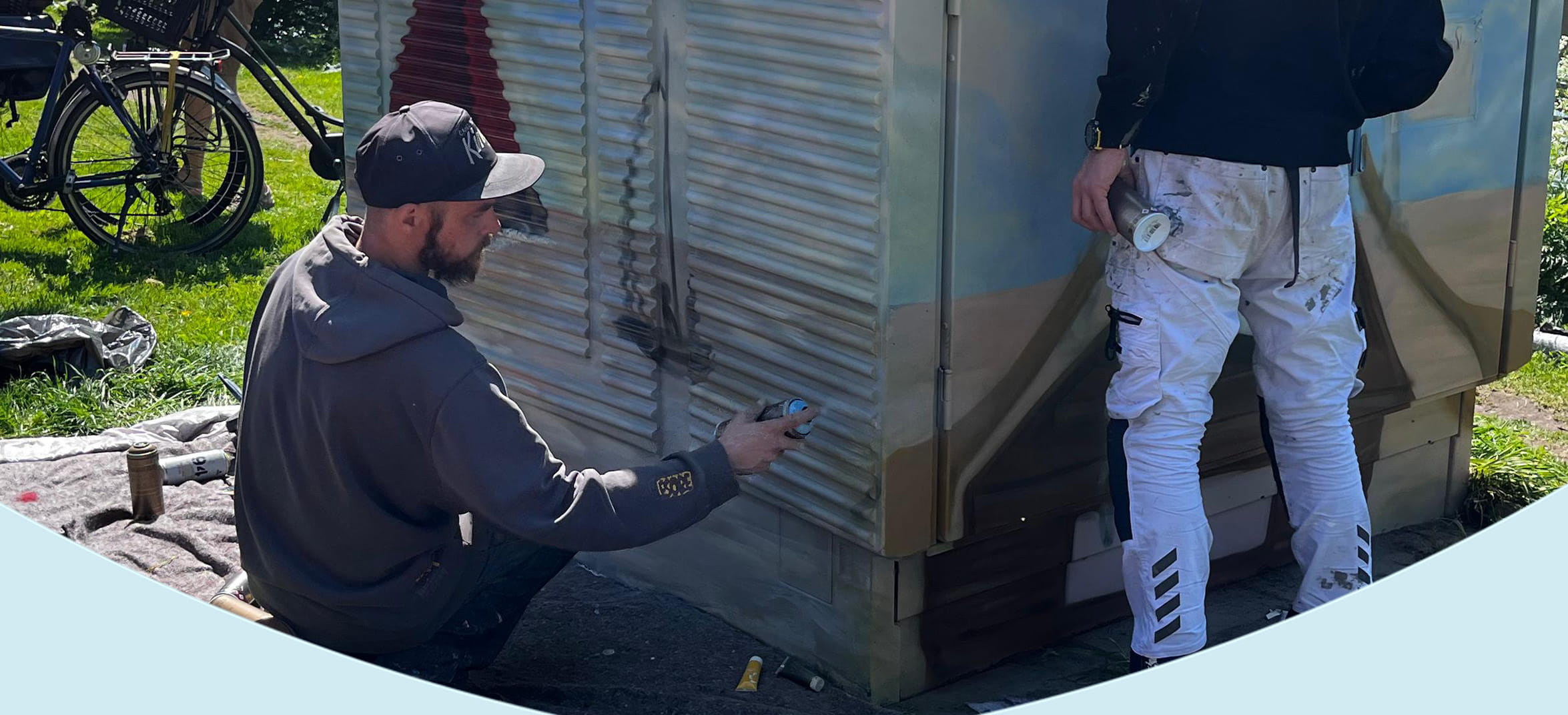 Twee kunstenaars spuiten graffiti op een elektriciteitshuisje langs een kanaal, omringd door verfspullen en toeschouwers in een parkachtige omgeving.