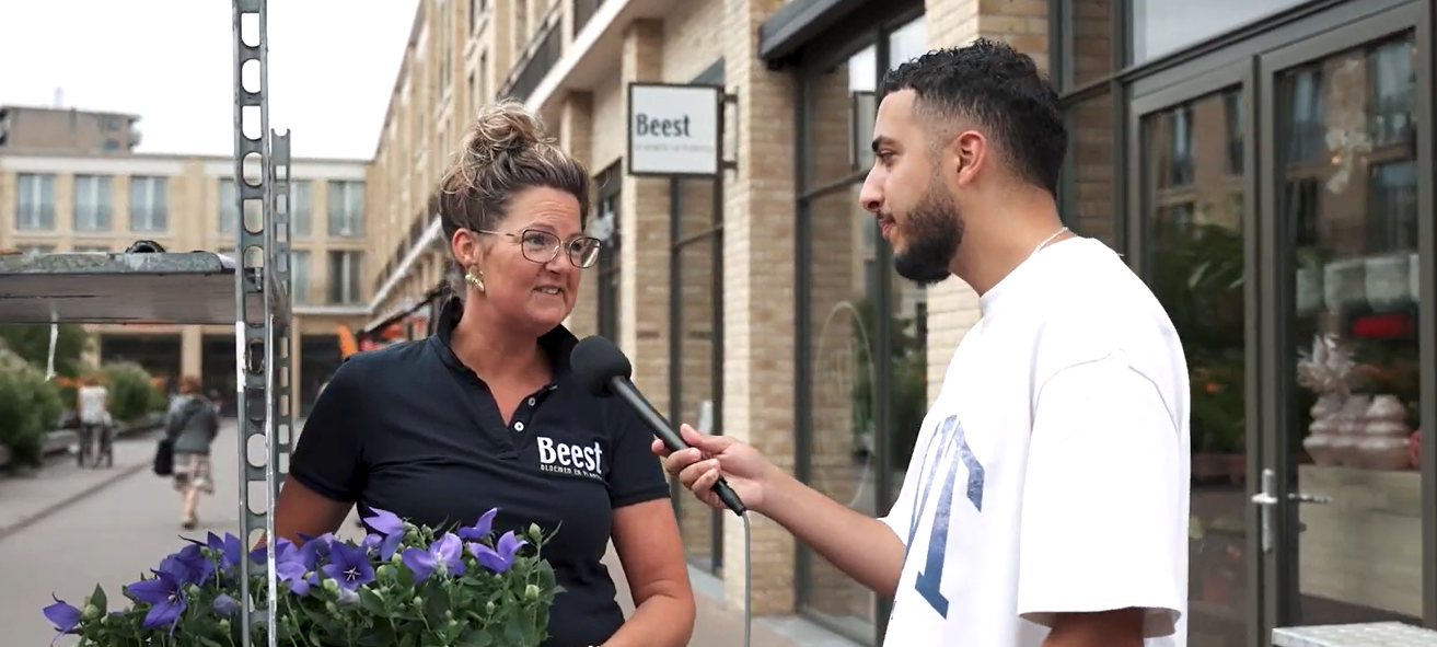 Een man houdt een microfoon vast en interviewt een vrouw met een bloempot op straat.