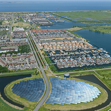 Luchtfoto van woonwijk in Almere. Op de voorgrond een zonnepark op een rond schiereiland. Op de achtergrond een groot meer