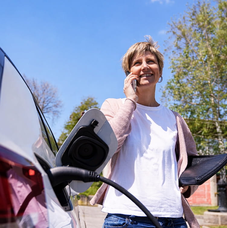 Een persoon staat naast een elektrische auto met een laadkabel in de hand. De auto is aangesloten op een laadpunt in een woonomgeving.