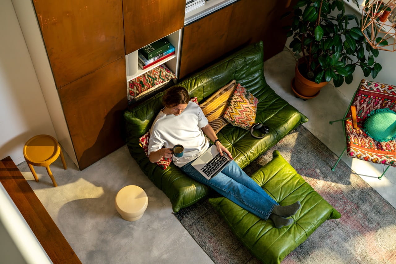 Persoon werkt met een laptop op een groene bank in een zonnige woonkamer met planten en kleurrijke kussens.
