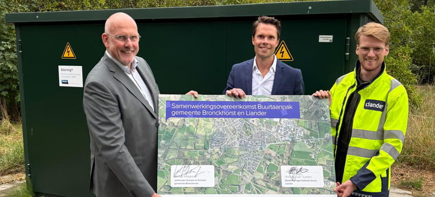 Drie mannen staan voor een donkergroen elektriciteitshuisje en houden een grote kaart vast met de tekst "Samenwerkingsovereenkomst Buurtaanpak gemeente Bronckhorst en Liander". Iedereen kijkt vrolijk in de camera