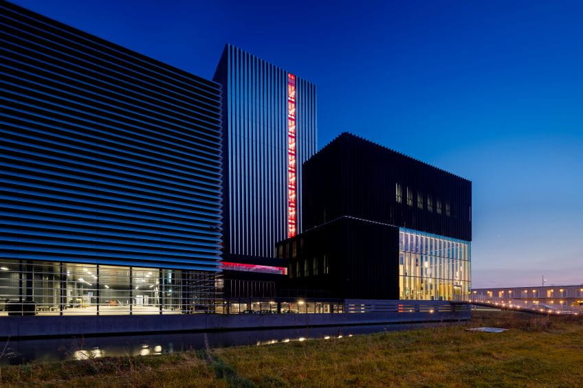 Meerdere moderne zwarte gebouwen die samen een datacenter vormen met glazen gevels en een rood verlichte toren, gefotografeerd bij avondlicht.