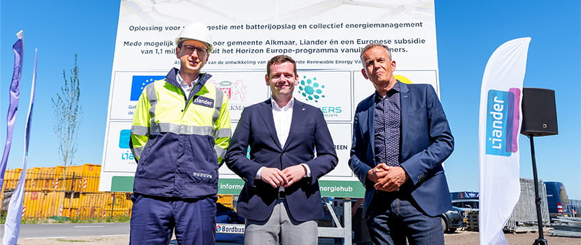 Drie mannen poseren voor een bouwbord van de Energiehub Boekelermeer in Alkmaar, die operationeel wordt in november 2025. Op het bord staat informatie over slimme stroomvoorziening, batterijopslag en Europese subsidie. Links staat een man in werkkleding van Liander met helm.