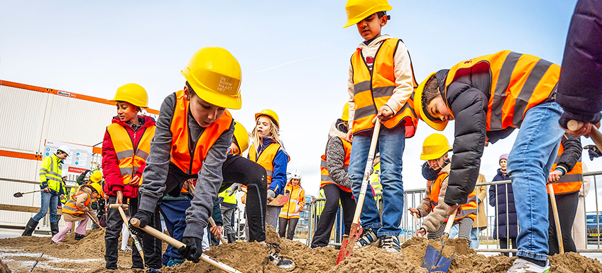 Kinderen staan met een schop bij een berg zand. Ze dragen een veiligheidshesje en helm. Ze zijn enthousiast aan het scheppen.