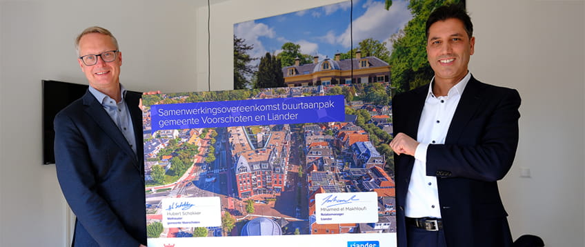 De Wethouder van Voorschoten en relatiemanager van Liander houden een groot bord vast met de tekst 'Samenwerkingsovereenkomst buurtaanpak gemeente Voorschoten en Liander', met een luchtfoto van een stadsgebied op de achtergrond van het bord. Ze staan glimlachend naast elkaar in een kantoorruimte.