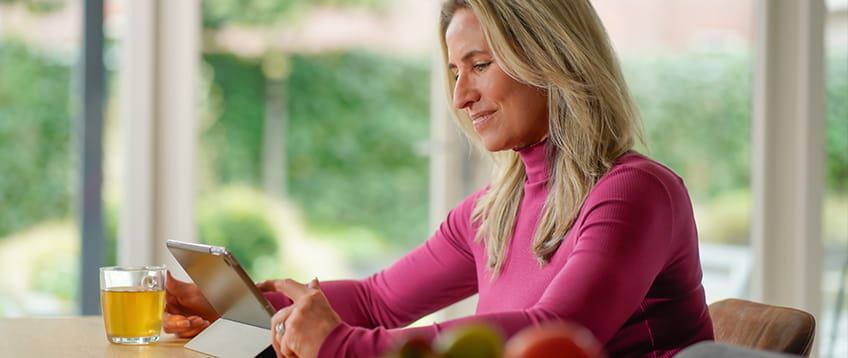 Blonde vrouw in roze trui kijkt op een Ipad die voor haar op tafel staat