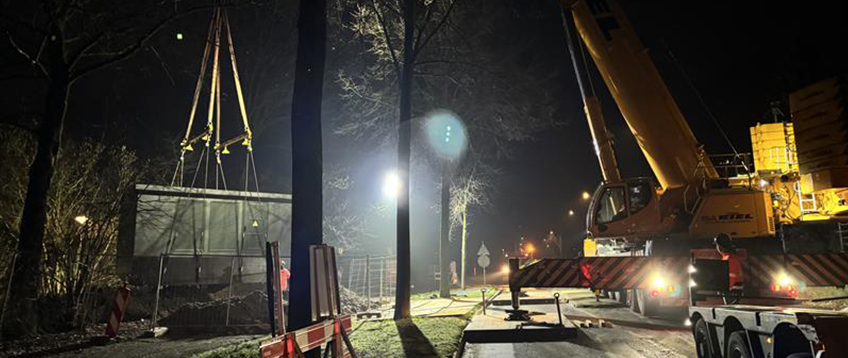 Gele hijskraan hijst een elektriciteitshuisje op zijn plek tijdens nachtelijke werkzaamheden. Het werk wordt verlicht door een grote spotlicht