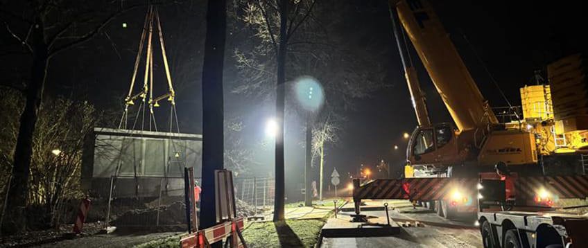 Gele hijskraan hijst een elektriciteitshuisje op zijn plek tijdens nachtelijke werkzaamheden. Het werk wordt verlicht door een grote spotlicht