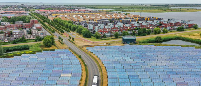 Luchtfoto van een groot veld met zonnepanelen langs een weg waar een bus rijdt, met daarachter een woonwijk en water.
