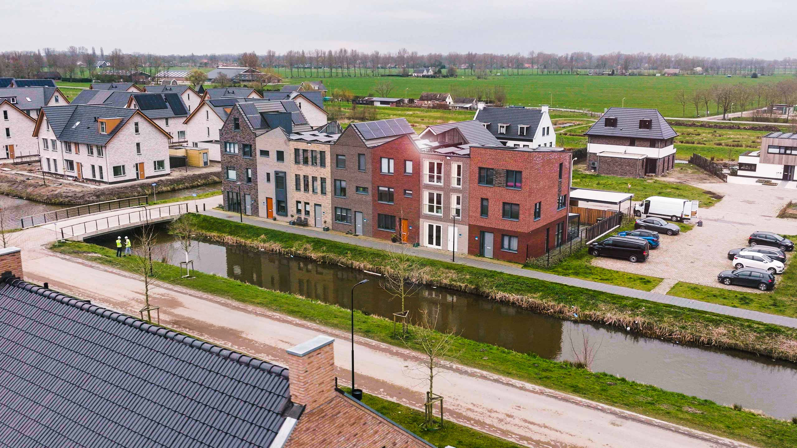 Luchtfoto van een nieuwbouwwijk met moderne rijtjeshuizen en zonnepanelen, langs een kanaal. Twee mensen in veiligheidshesjes staan op een zandpad bij het water.
