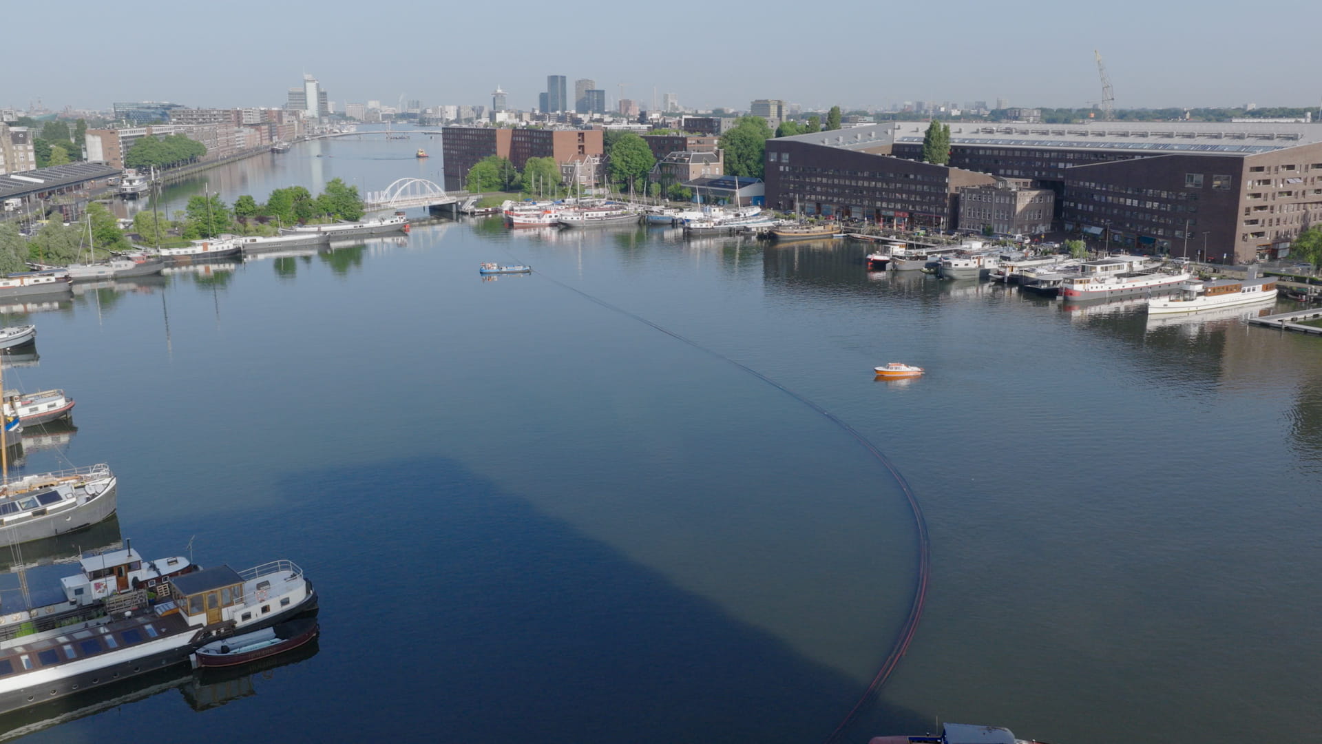 Luchtfoto van Sporenburg Amsterdam. Je ziet een rustige haven met verschillende boten aan de kades en een klein oranje bootje op het water. Op de achtergrond zijn moderne gebouwen en bruggen zichtbaar onder een heldere blauwe hemel.