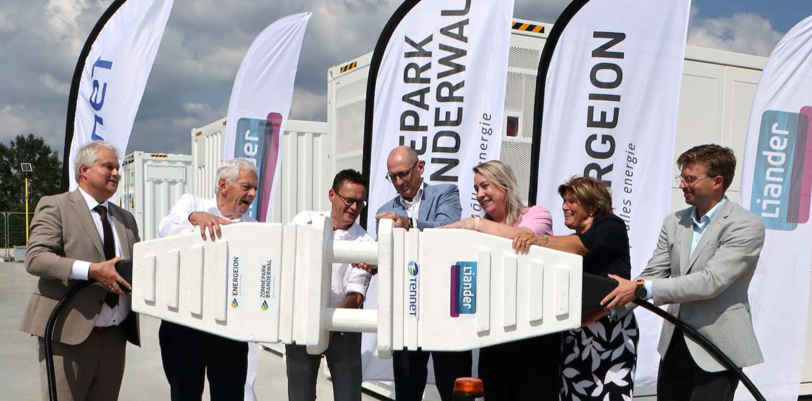 Meerdere personen koppelen symbolisch twee grote stekkers aan elkaar tijdens de opening van een energieproject, met energiecontainers en vlaggen van Liander en TenneT op de achtergrond.