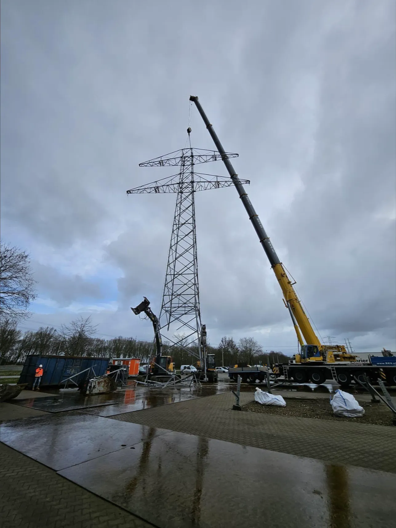 Mobiele kraan ondersteunt een hoogspanningsmast terwijl deze in delen wordt gedemonteerd.
