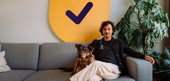 Een man zit op een bank met een hond naast zich, met een groot geel bord met een vinkje op de achtergrond.