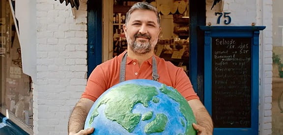 Een glimlachende man met een schort houdt een grote wereldbol vast voor een winkel.