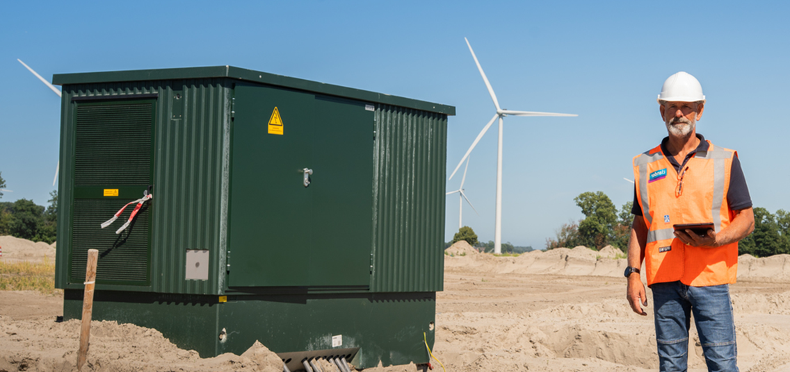 Persoon naast groene elektrische apparatuur met windmolens op de achtergrond.