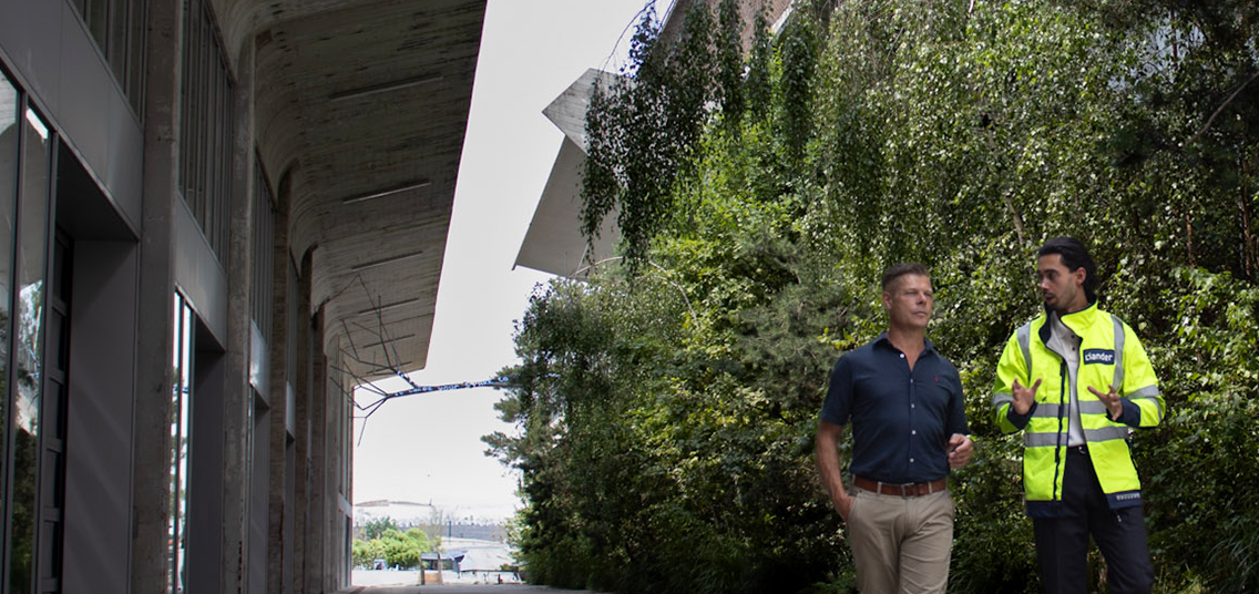 Twee personen wandelen en praten bij een gebouw met groene beplanting.