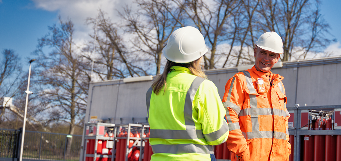 Twee medewerkers in veiligheidskleding in gesprek bij technische apparatuur.