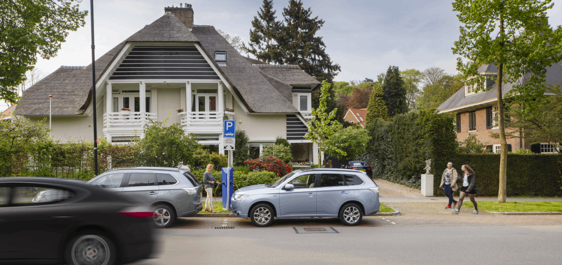 Grote witte vrijstaande woning, met grijs dak. Op de straat voor het huis staan twee grijze auto's geparkeerd bij een laadpaal.