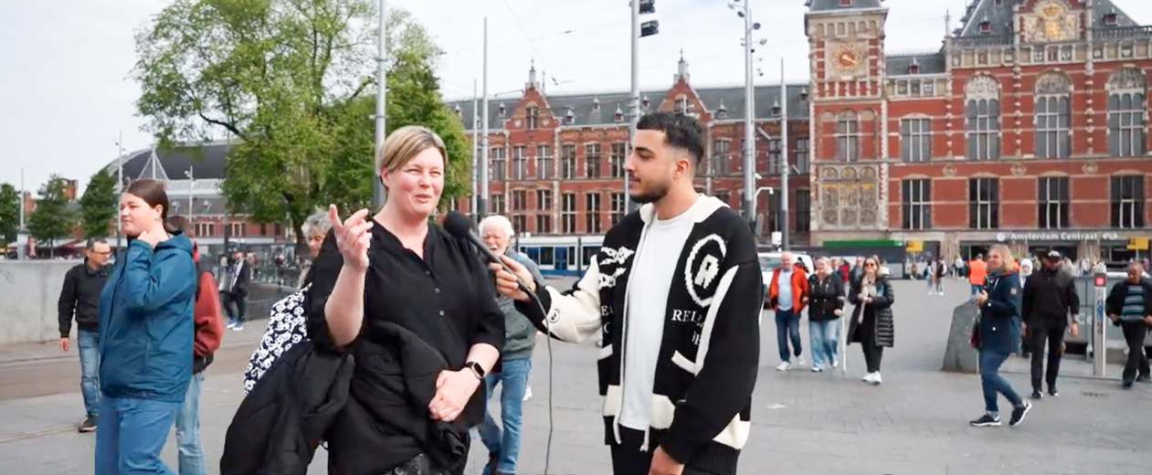 Straatinterview op een levendig plein bij Amsterdam Centraal Station, waar een interviewer een vrouw spreekt te midden van voorbijgangers en historische architectuur.