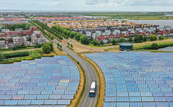 Luchtfoto van een landschap met grote zonnepaneelvelden aan weerszijden van een weg waar een bus rijdt, met op de achtergrond een woonwijk omringd door groen.